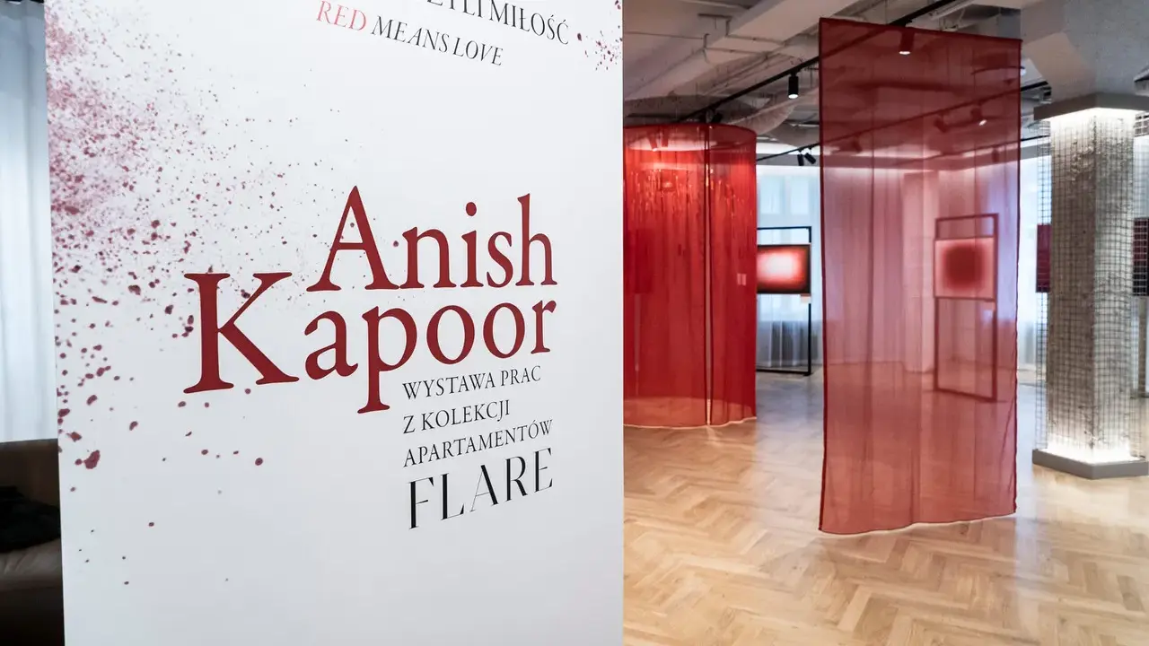 Biały plakat z napisem "Anish Kapoor" i czerwonymi rozpryskami na tle wystawy z czerwonymi zasłonami.