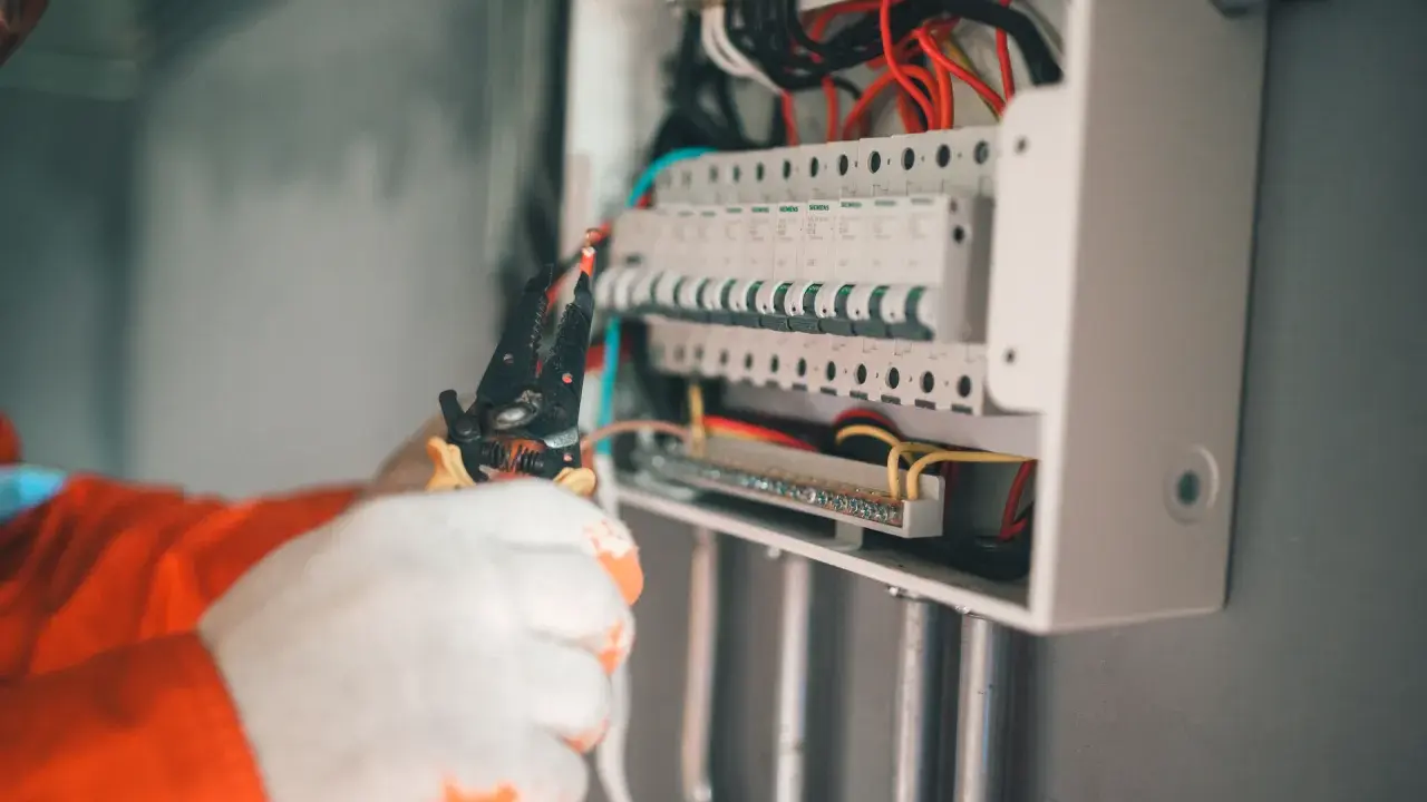 Elektryk w białych rękawicach manipuluje przewodami w skrzynce z bezpiecznikami. Wybija bezpiecznik bez obciążenia, co może sugerować problem.
