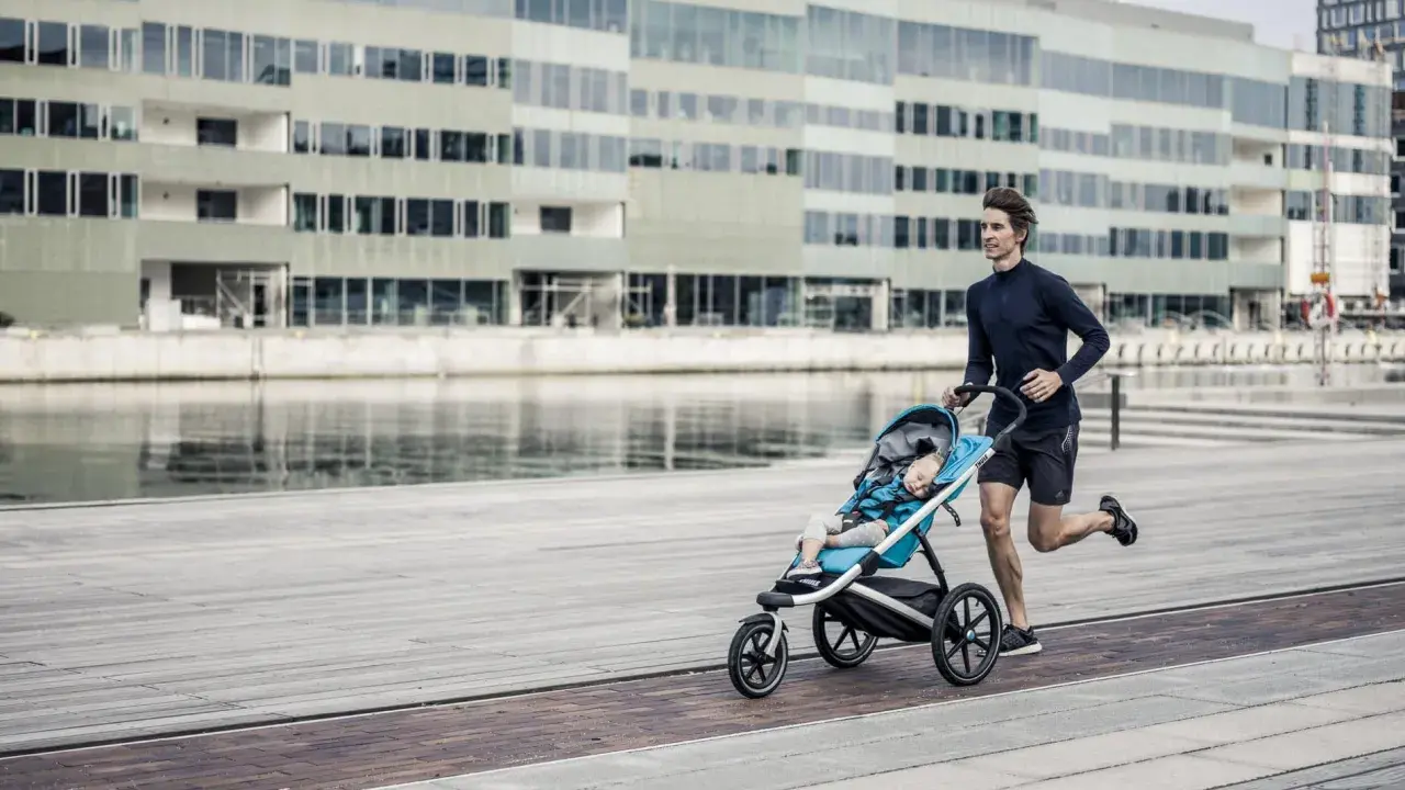 Ein Mann joggt mit Kinderwagen an einer modernen Fassade entlang. Das Kind schläft friedlich im Wagen.