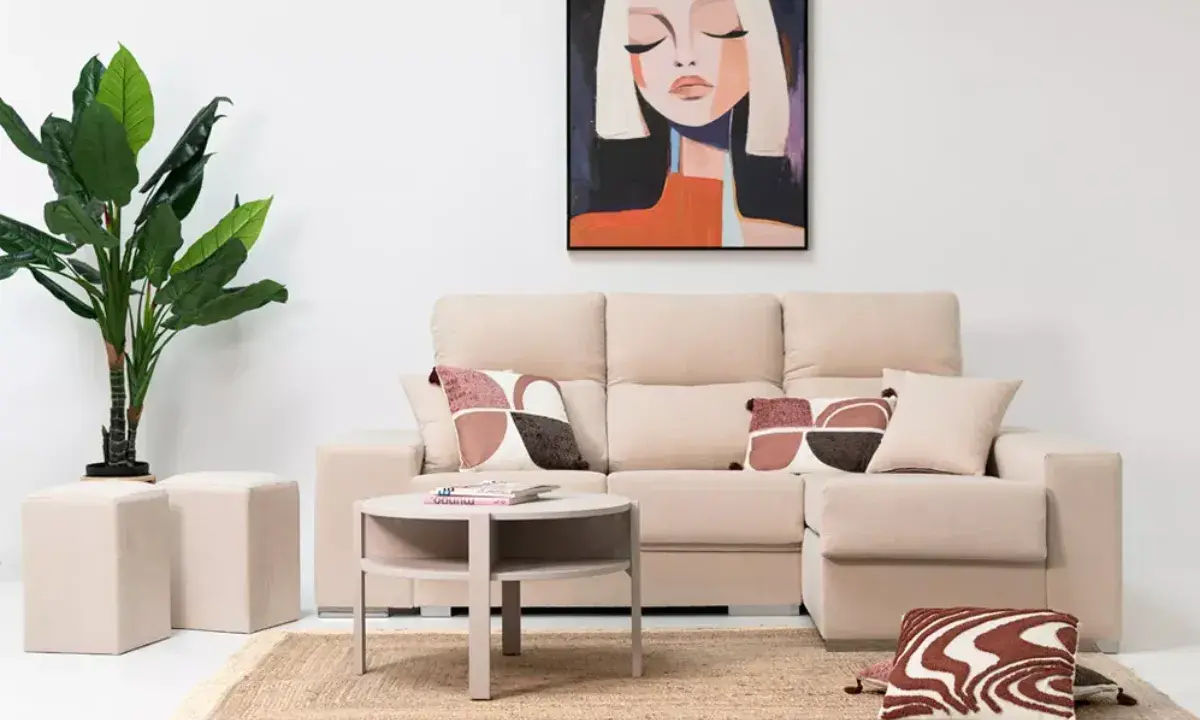 Sofá modular beige con chaise longue a la derecha, mesa auxiliar redonda, alfombra de yute, plantas y cuadro abstracto.