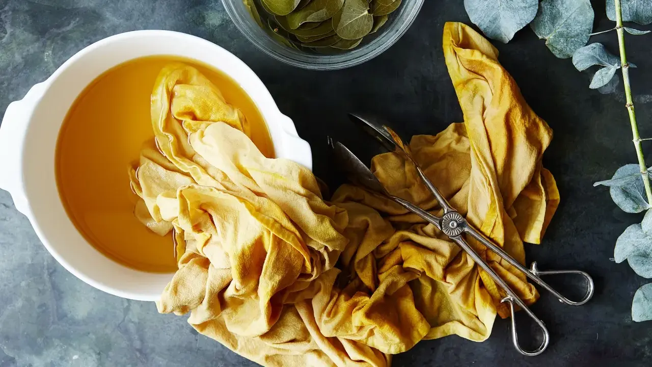 A yellow fabric is soaking in a bowl of golden liquid, part of a natural dye process. Eucalyptus leaves are nearby.