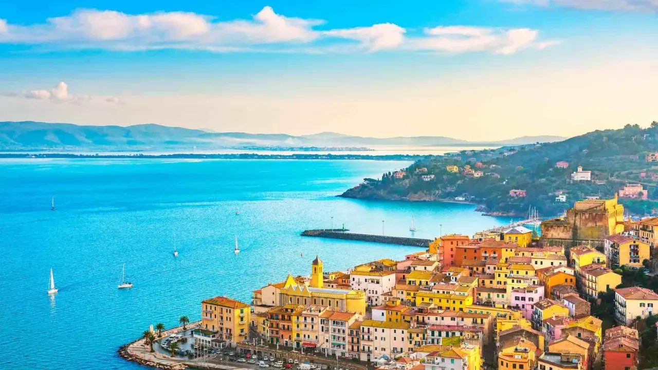 Colorate case affacciate sui mari della Toscana, con barche a vela che solcano le acque azzurre sotto un cielo limpido.
