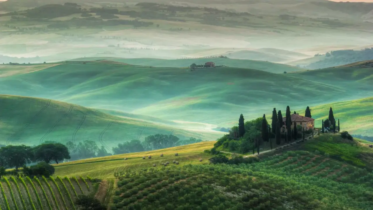 Un incantevole **punto panoramico Val d'Orcia** con dolci colline verdi, un casale in cima a una collina e filari di cipressi.