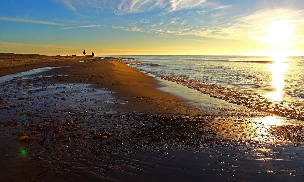 Två personer promenerar längs stranden vid Skagens udde i solnedgången. Vågorna sköljer in mot land.
