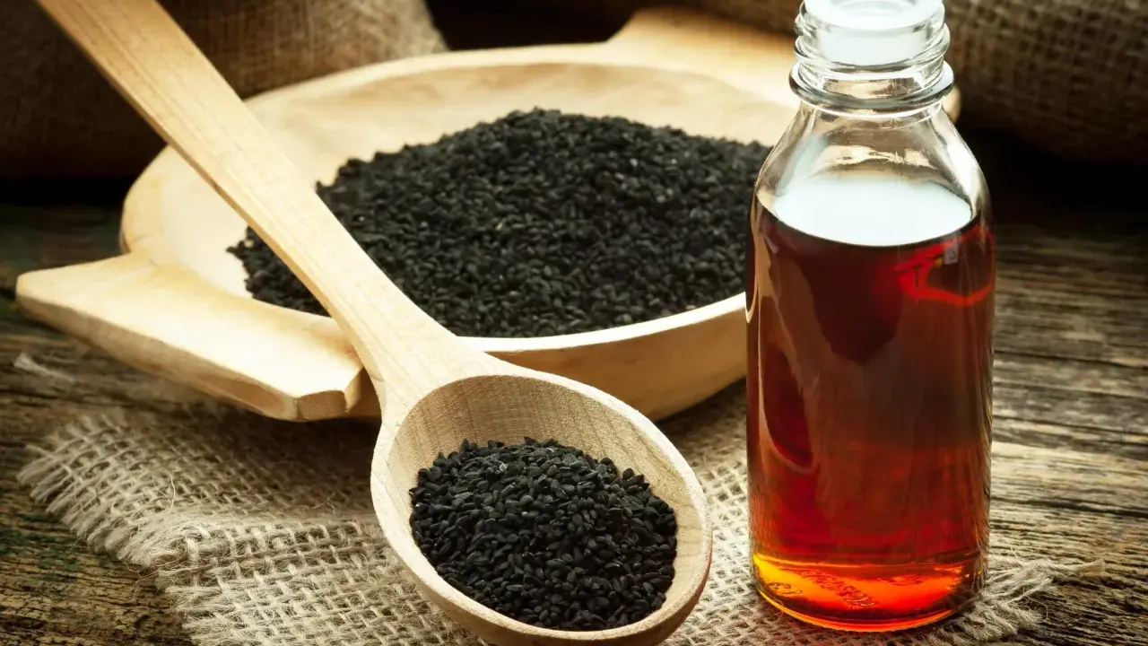 Huile de nigelle ambrée dans un flacon, à côté de graines noires dans une cuillère en bois. Découvrez les bienfaits de l'huile de nigelle.