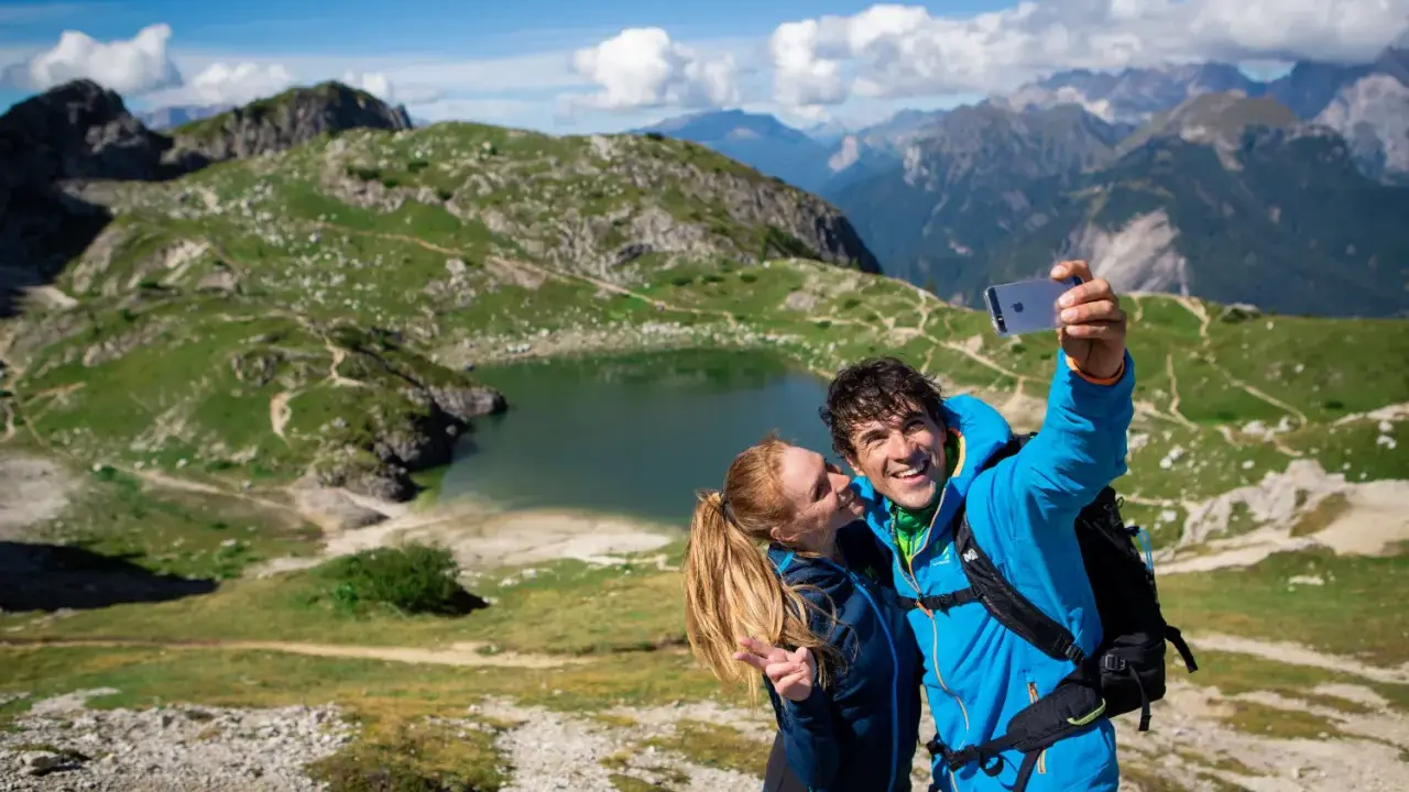 Coppia felice si scatta un selfie con il lago Coldai sullo sfondo, circondato da montagne verdi e un cielo azzurro con nuvole bianche.