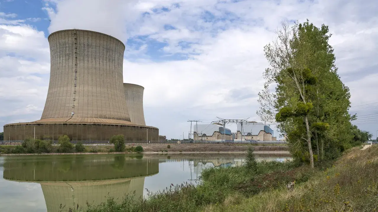 Kühltürme eines Atomkraftwerks in Frankreich, die Dampf abgeben. Ein ruhiger See spiegelt die Anlage wider, während am Ufer Bäume und Gras wachsen.