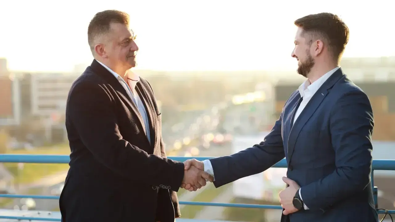 Dwóch mężczyzn w garniturach ściska sobie dłonie na tle miasta.