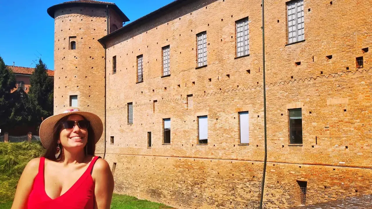 Donna sorridente con cappello di paglia davanti a un castello di mattoni rossi. Un'idea per cosa vedere a Piacenza in un giorno.