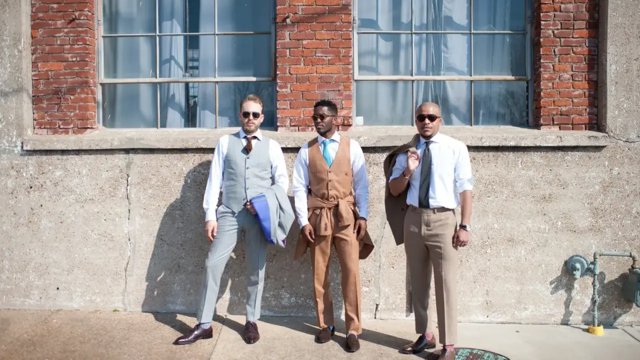Three men in stylish suits embody cocktail chic attire. One wears a grey pinstripe suit, another a tan suit with a turquoise tie, and the third a light blue shirt with tan trousers.