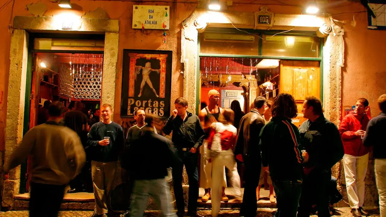 Menschen versammeln sich vor einem beleuchteten Lokal in Bairro Alto. Ein Mann hält ein Glas, andere unterhalten sich.