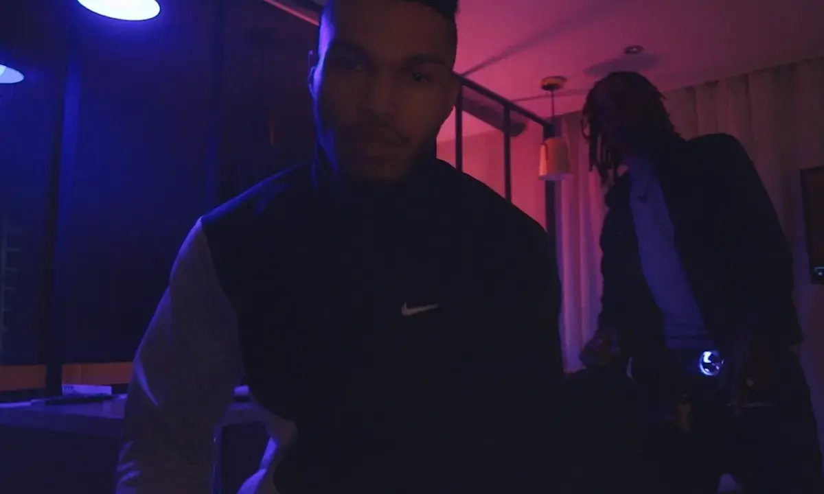 Dwóch mężczyzn w ciemnym pomieszczeniu, oświetlonym niebieskim i różowym światłem. Jeden z nich, 730 huncho, ma na sobie bluzę z logo Nike.