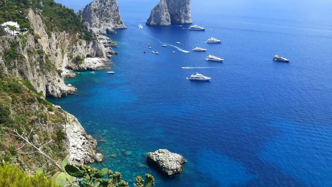 Scogliere verdi e rocce imponenti si affacciano sul mare blu intenso, con yacht che navigano. Un'immagine che ispira su come raggiungere Capri da Napoli.