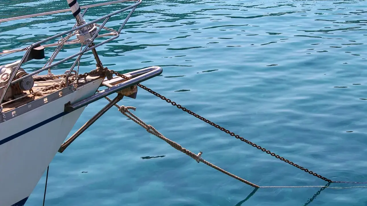 L'étrave d'un voilier blanc est ancrée dans une eau bleue et claire. La chaîne et la corde sont bien visibles, prêtes à mouiller beaucoup.
