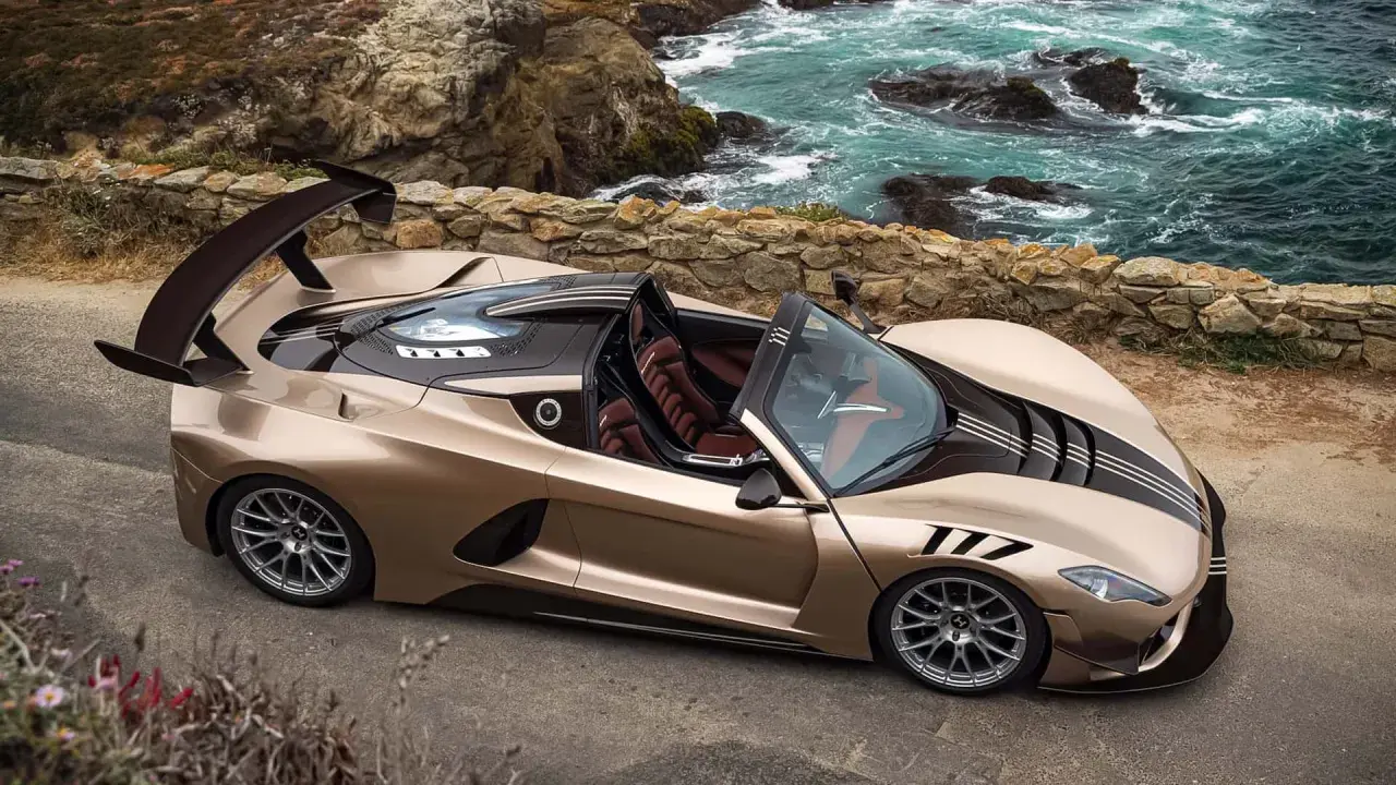 Der Hennessey Venom F5 Roadster in Goldbraun steht an einer felsigen Küste. Sein aggressives Design mit großem Heckspoiler und dunklen Streifen zieht alle Blicke auf sich.