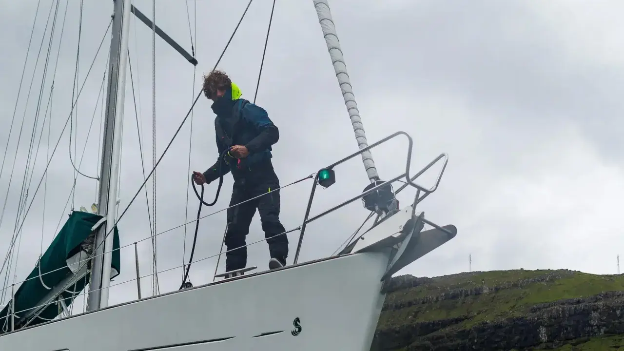 Un marinier, les amarres avis, manœuvre sur le pont d'un voilier blanc, face à une côte rocheuse verdoyante.