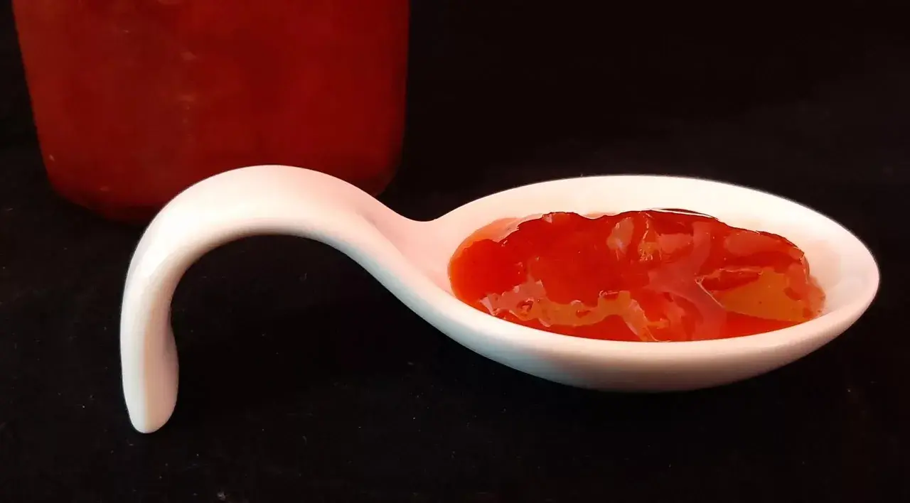 Une cuillère blanche présente une portion de confiture fraise rhubarbe, d'un rouge vif et appétissant. Un pot de confiture est visible en arrière-plan.