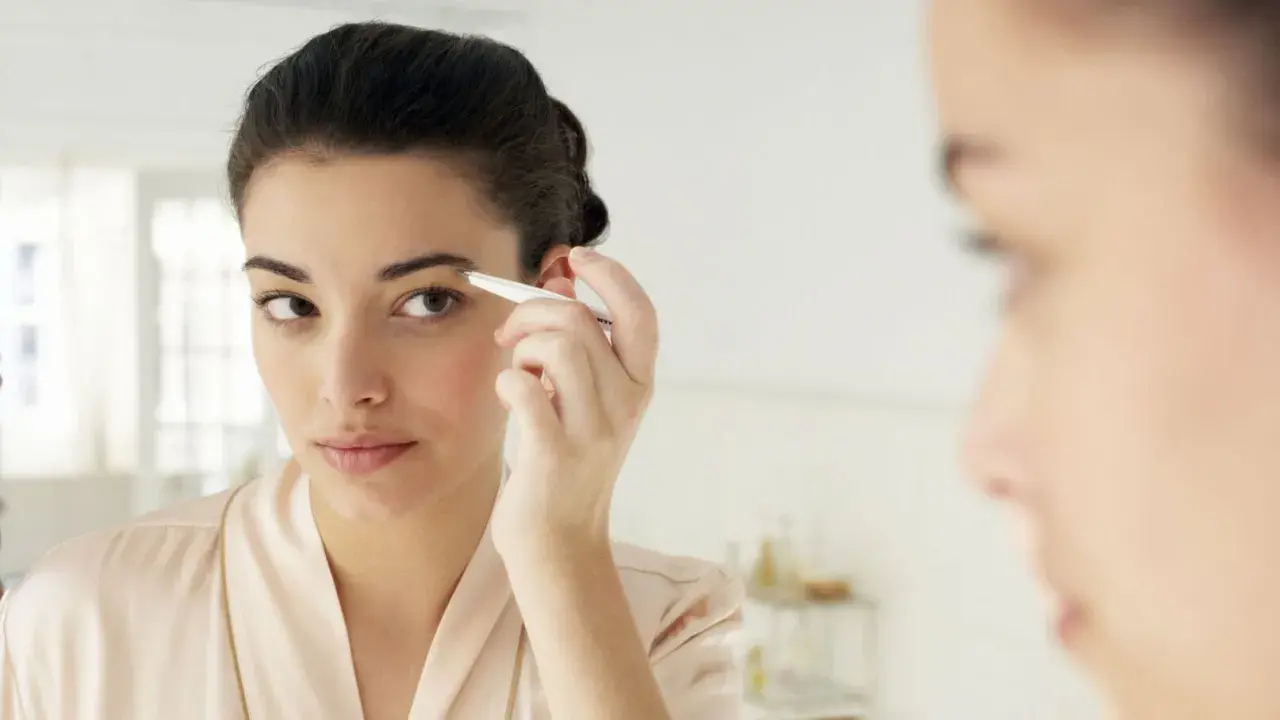 Une jeune femme ajuste ses sourcils broussailleux avec une pince à épiler devant un miroir, dans une tenue de chambre satinée.