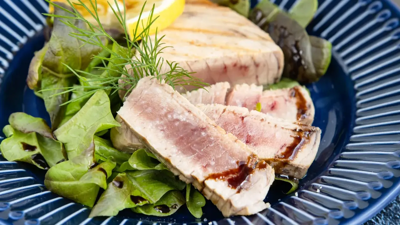 Ein perfekt gegrilltes Thunfischsteak, in Scheiben geschnitten, serviert mit frischem Salat, Zitrone und Dill auf einem blauen Teller.