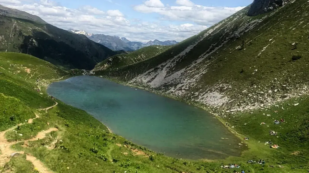 Un sentiero serpeggia tra prati verdi verso il lago Branchino, le cui acque turchesi riflettono le montagne circostanti. Alcune persone si godono un picnic sulle rive.