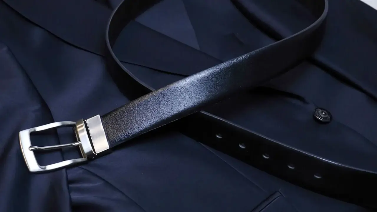 A black leather belt with a silver buckle rests on a dark navy suit jacket. Do you wear a belt with a tuxedo? This image shows a classic accessory.