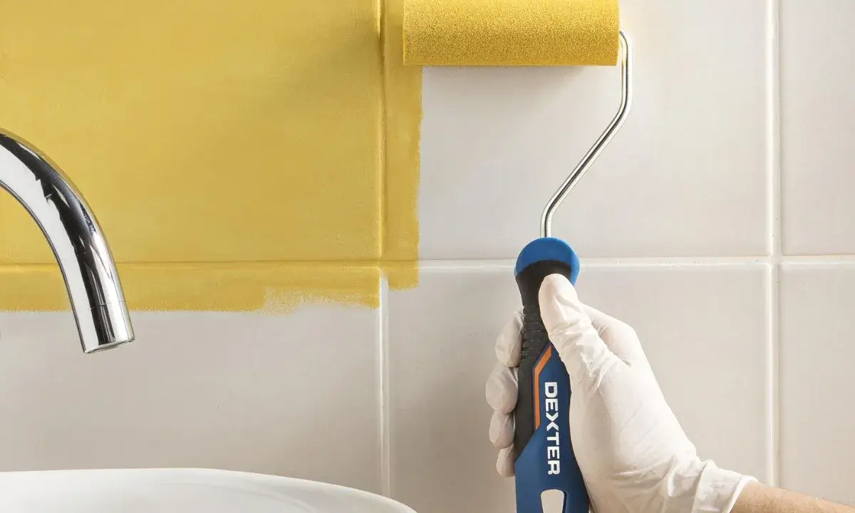 Mano con guante blanco pintando azulejos de cocina de blanco a amarillo con rodillo.