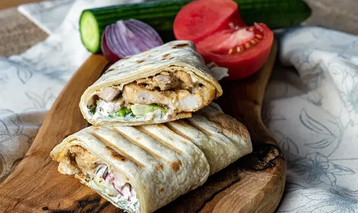 Pyszny, domowy kebab z kurczakiem, warzywami i sosem na desce z drewna. Obok pomidor, ogórek i cebula.