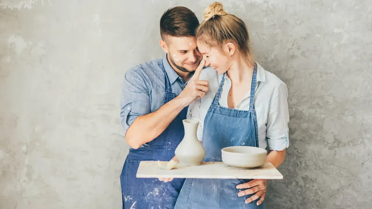 Para świętuje 9 rocznicę ślubu, tworząc ceramikę. Mężczyzna delikatnie dotyka nosa kobiety, oboje ubrani w fartuchy, ubrudzeni gliną.
