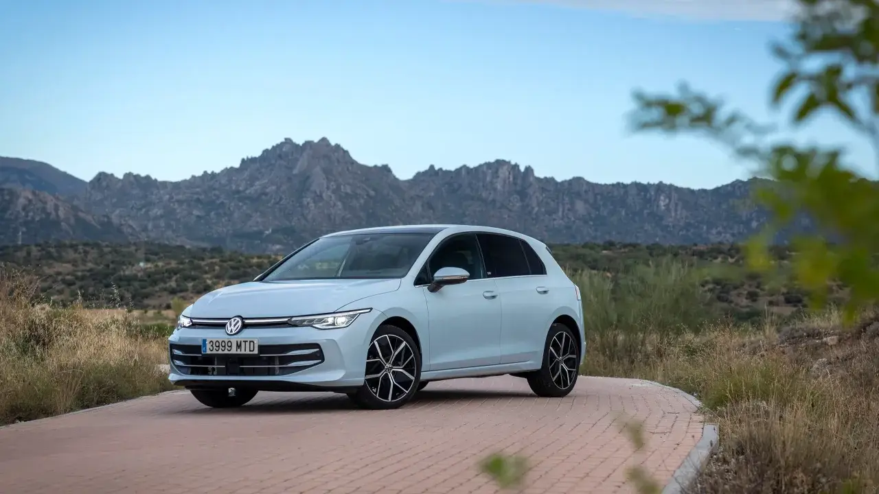 Un coche compacto Volkswagen Golf blanco circula por una carretera de adoquines con montañas rocosas al fondo.