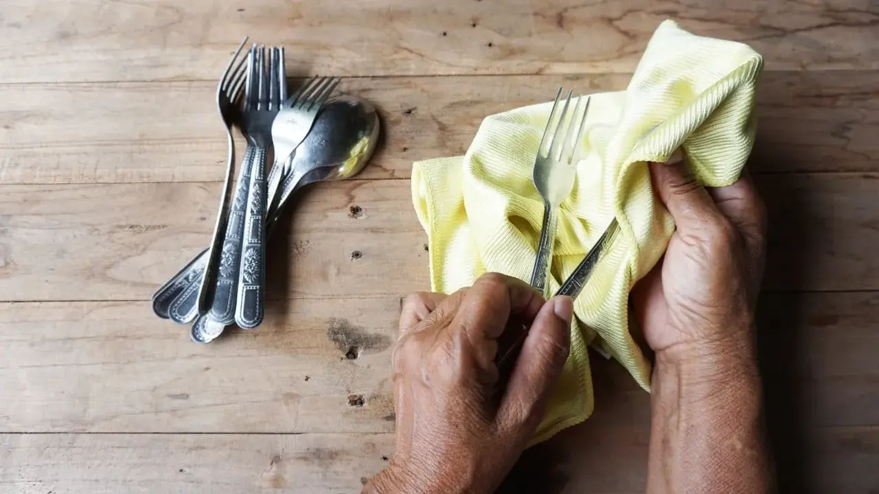 Mains polissant des couverts en argent avec un chiffon jaune. Un tas de fourchettes et cuillères est à gauche. Apprenez comment nettoyer l'argenterie.