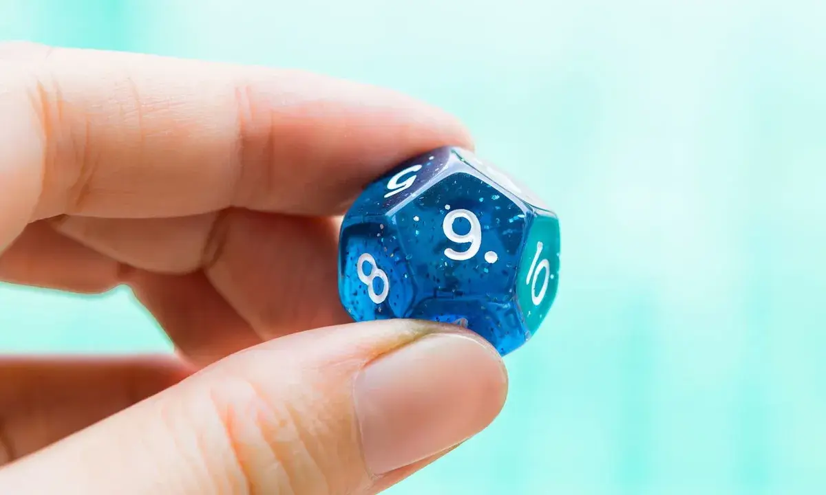Palec i kciuk trzymają niebieską, dwudziestościenną kostkę z błyszczącymi drobinkami. Widoczna jest cyfra 9, symbolizująca numerologiczną 9.