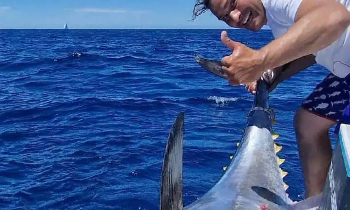 Uomo felice mostra il suo **biggest tuna** appena pescato in mare aperto, con una barca e una vela all'orizzonte.