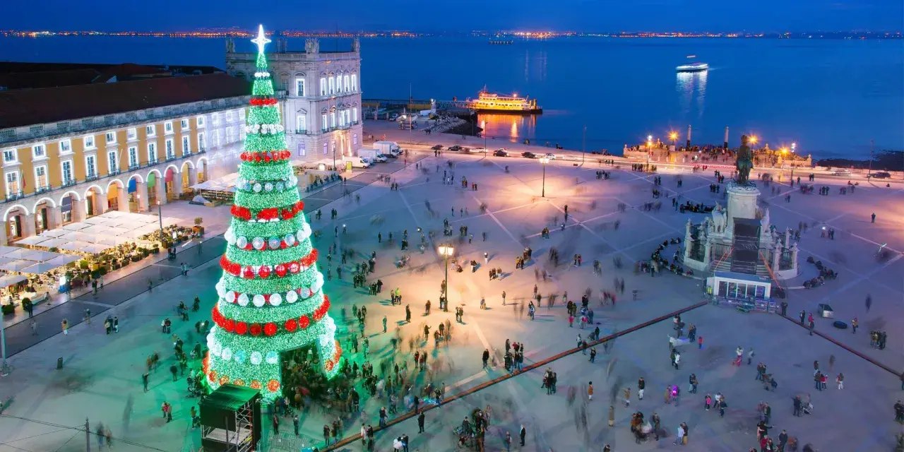 Lisbona illuminata per il weekend dell'Immacolata: un imponente albero di Natale brilla in una piazza affollata, con il fiume e le luci della città sullo sfondo.
