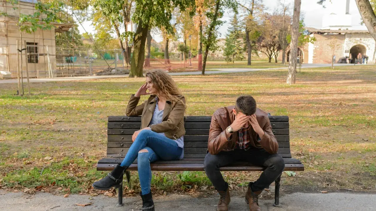 Pareja sentada en un banco, ella mira a un lado, él se tapa la cara. Parecen estar intentando procesar amor y desamor en un parque otoñal.
