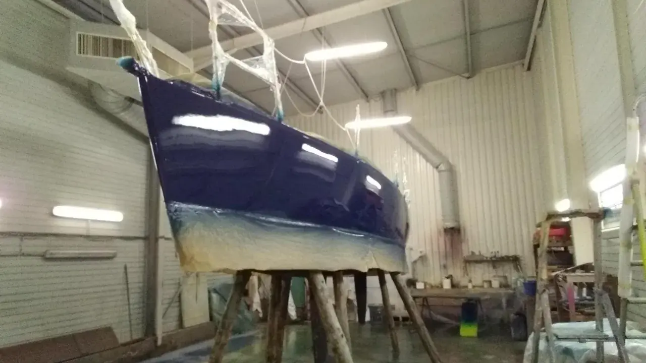 Un bateau bleu profond est en cours de stratification dans un atelier. La coque est soutenue par des tréteaux en bois.