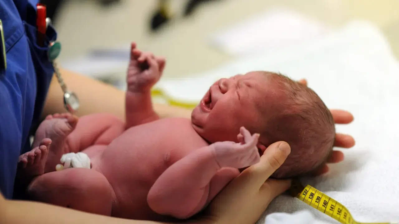 Neugeborenes Baby schreit, die Kopfform ist gut sichtbar. Ein Maßband liegt um den Kopf, wie nach einer Geburt mit Saugglocke.