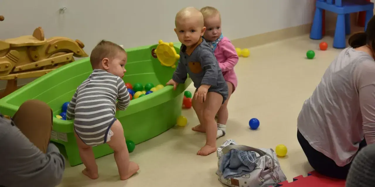 Trzy maluchy, w tym 6 miesięczne dziecko w chodziku, bawią się kolorowymi piłeczkami w zielonej wanience.