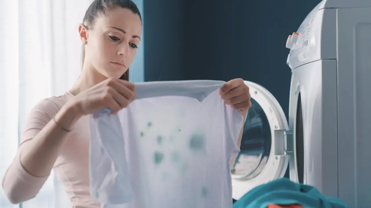 Zmartwiona kobieta trzyma białą koszulkę z zielonymi plamami, która wygląda jak zafarbowane ubrania. Obok stoi otwarta pralka.