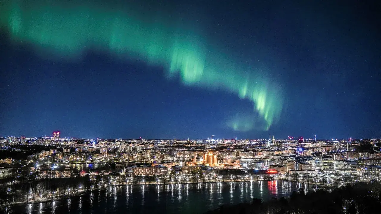 Grön norrskensbåge lyser upp natthimlen över en stad vid vattnet. En vacker demonstration av hur bildas norrsken.