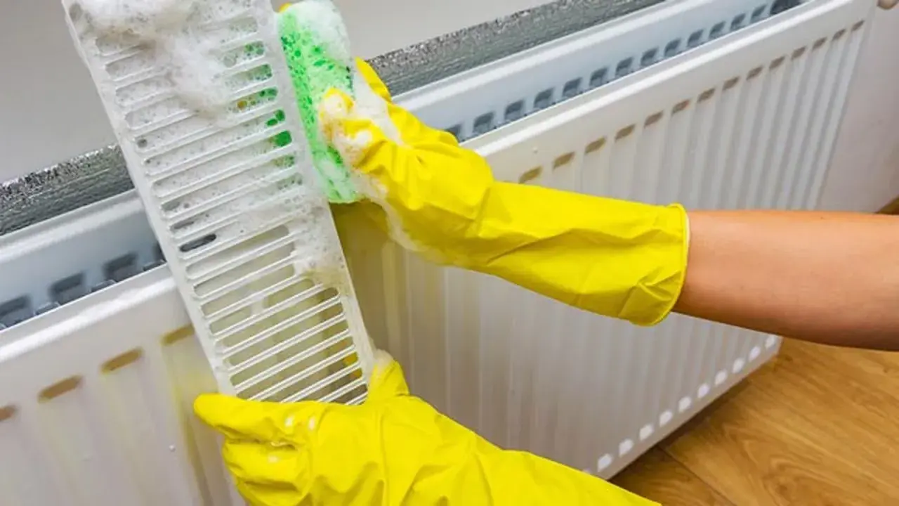 Manos con guantes amarillos limpian la rejilla de un radiador con una esponja y espuma.