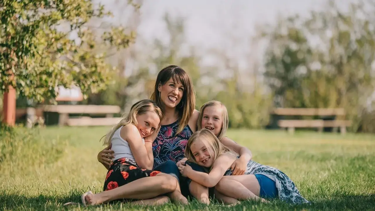 Mama z córkami na trawie, uśmiechnięte i przytulone. Widać, że czują motyle w brzuchu od tej miłości.