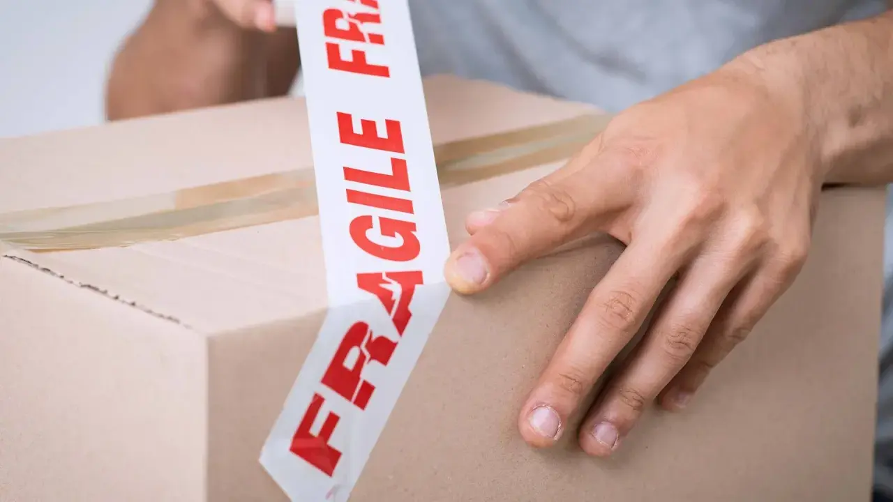 Ręce zaklejają karton taśmą z napisem "FRAGILE". Uwaga szkło, kurier musi być ostrożny.