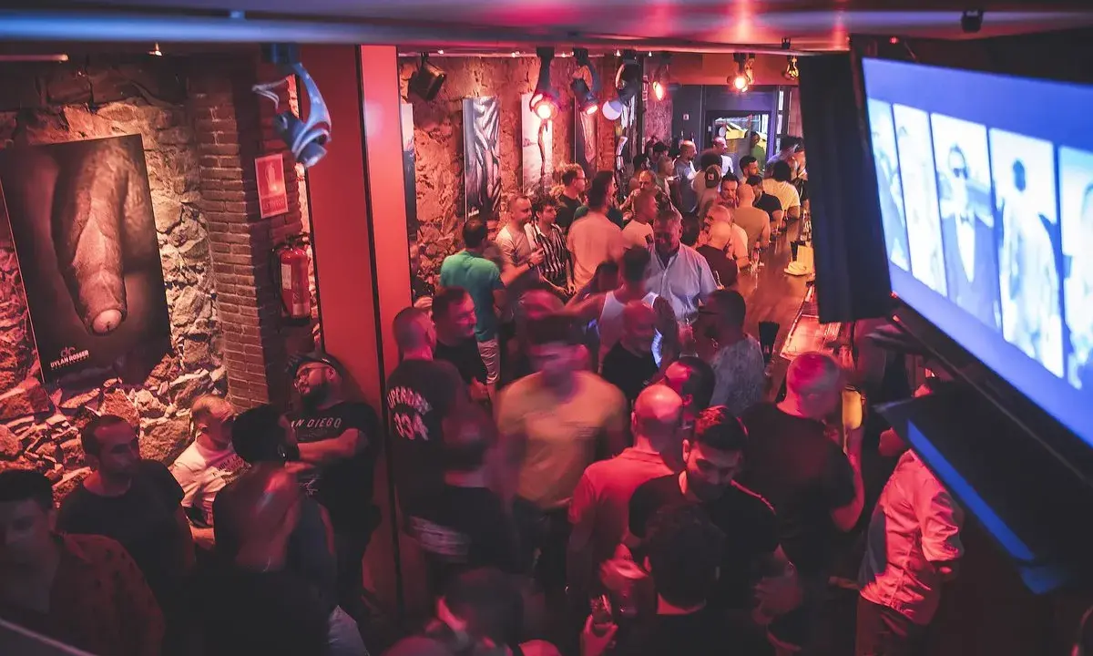 Un locale affollato a Barcellona by night, con uomini che socializzano e bevono. L'atmosfera è vibrante, illuminata da luci rosse e blu.