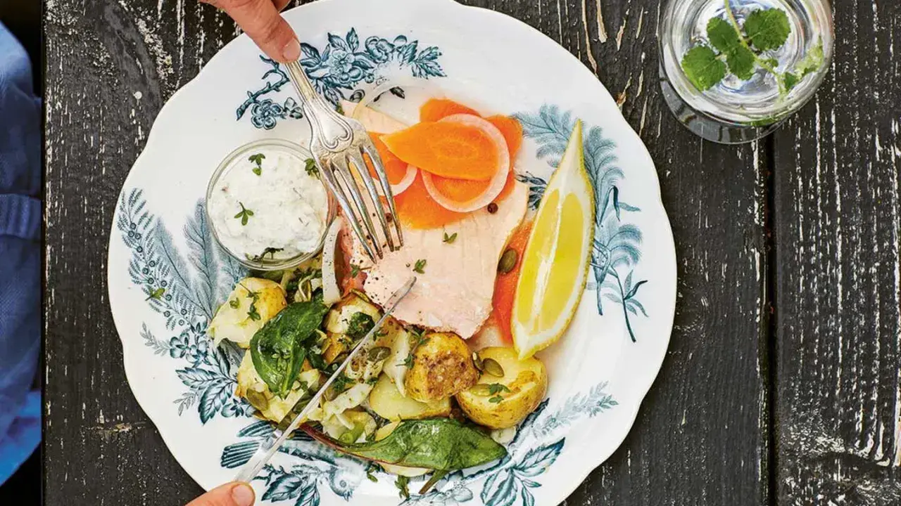 En tallrik med svensk lax, potatis, grönsaker och en citronskiva. En person skär i laxen med en gaffel och kniv.