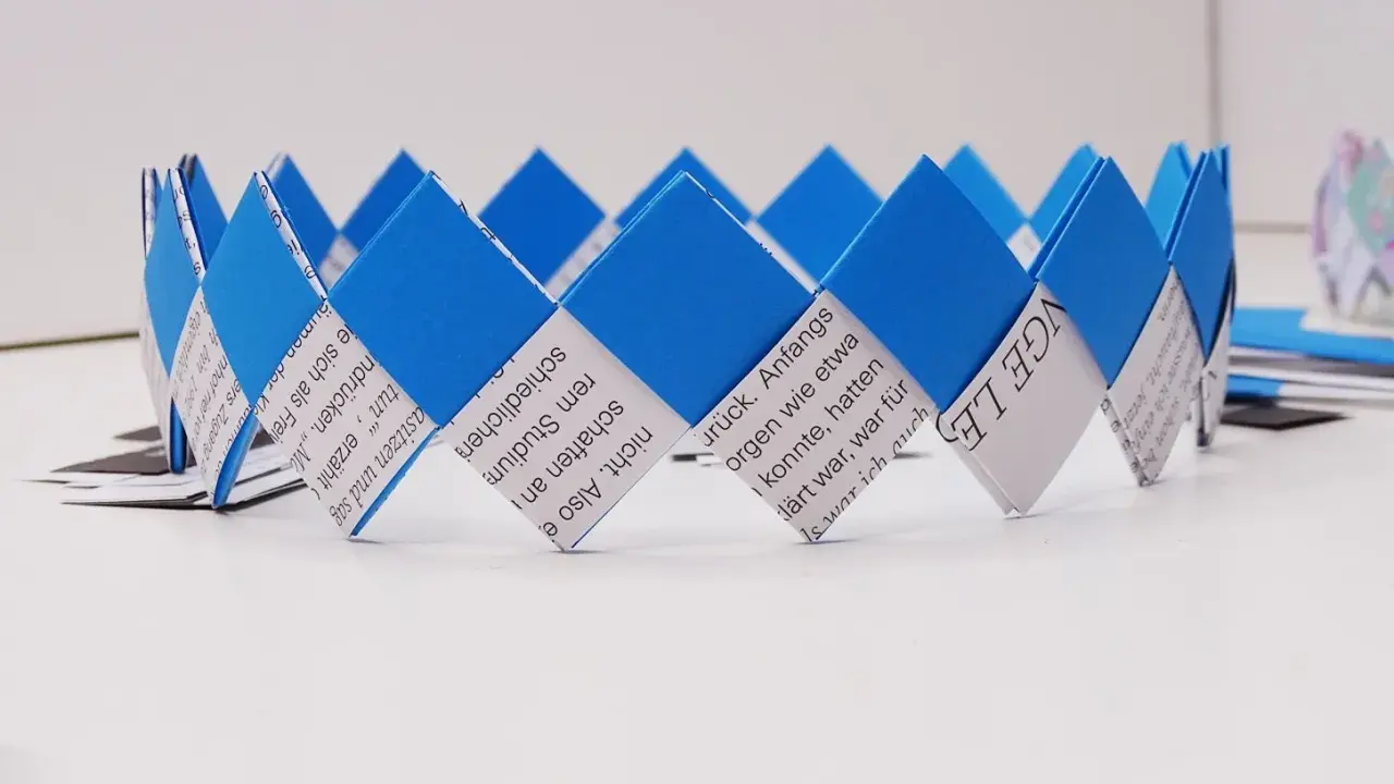 Eine blaue und weiße Papierkrone, die aus gefalteten Papierstreifen gefertigt wurde. Die Streifen sind mit Text bedruckt, was die Krone zu einem besonderen Kunstwerk macht.
