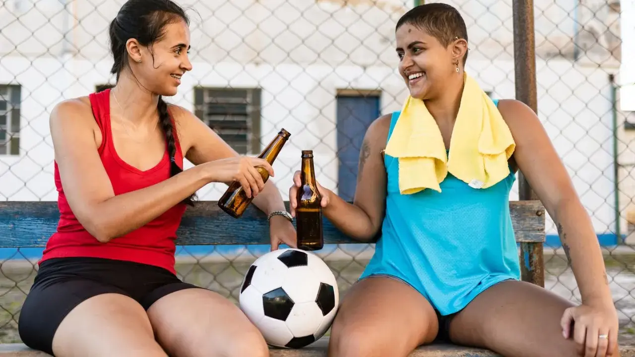 Zwei Frauen mit Fußball und Bierflaschen stoßen an. Nach dem Sport ist vor dem Genuss: sport und alkohol.
