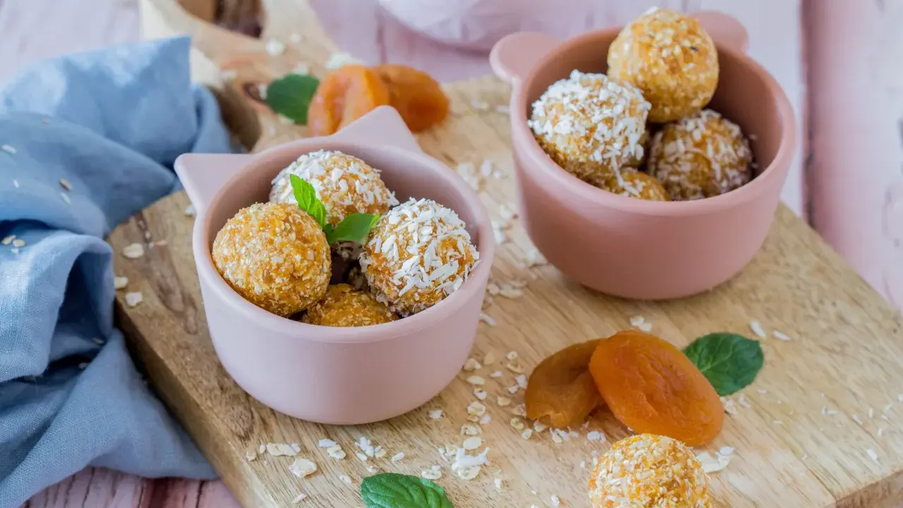 Zwei rosa Schalen gefüllt mit leckeren Energy Balls, bestreut mit Kokosraspeln und Haferflocken. Getrocknete Aprikosen und Minzblätter liegen daneben.
