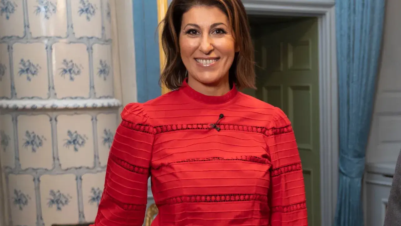 Rania Shemoun Olsson ler i en röd blus med puffärmar. Bakom henne syns en kakelugn och en dörröppning.