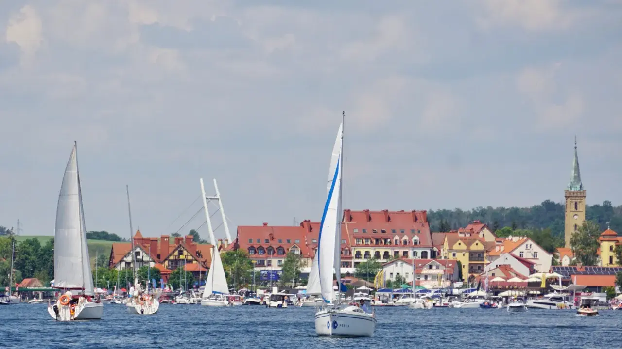 Piękne mazury widoki: żaglówki na jeziorze, miasteczko z czerwonymi dachami i wieżą kościelną w tle.