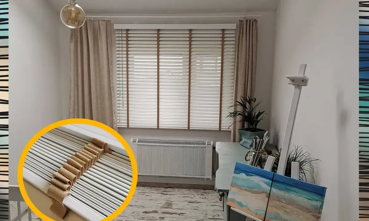 Jasne okna z żaluzjami w środku, ozdobione sznurkiem, tworzą przytulną atmosferę. Obok sztaluga z obrazem plaży.